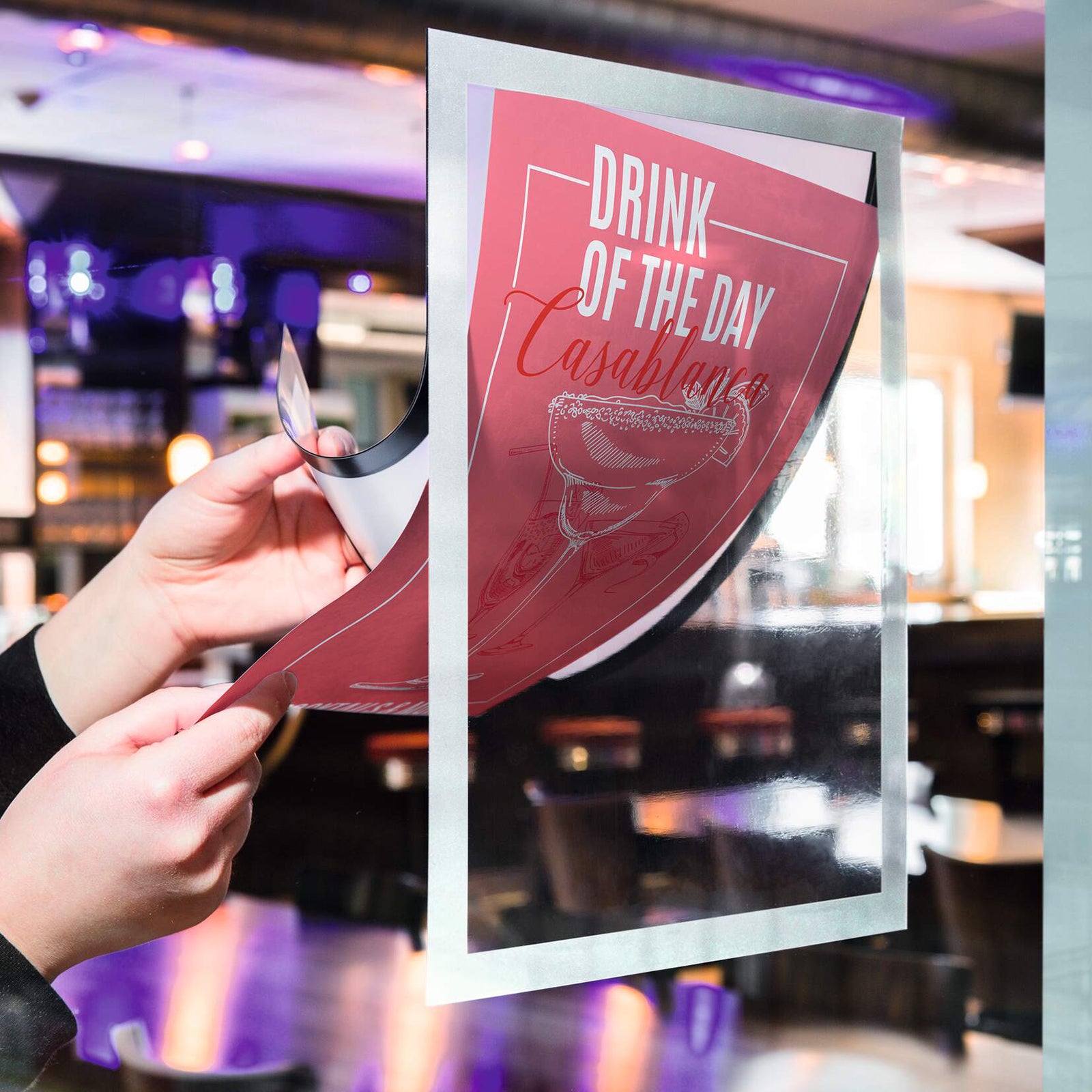 Hands updating a promotional drink poster inside the Durable DURAFRAME Sun A4 Silver on a glass surface in a hospitality setting. The fold back magnetic frame makes document changes quick while maintaining a professional display.