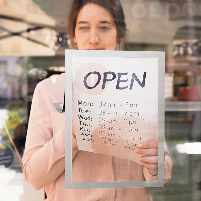 Retail staff member placing the Durable DURAFRAME Sun A4 Silver on a glass storefront to display opening hours. The image shows the clean silver frame, clear document visibility and static cling attachment suited to customer facing business signage.
