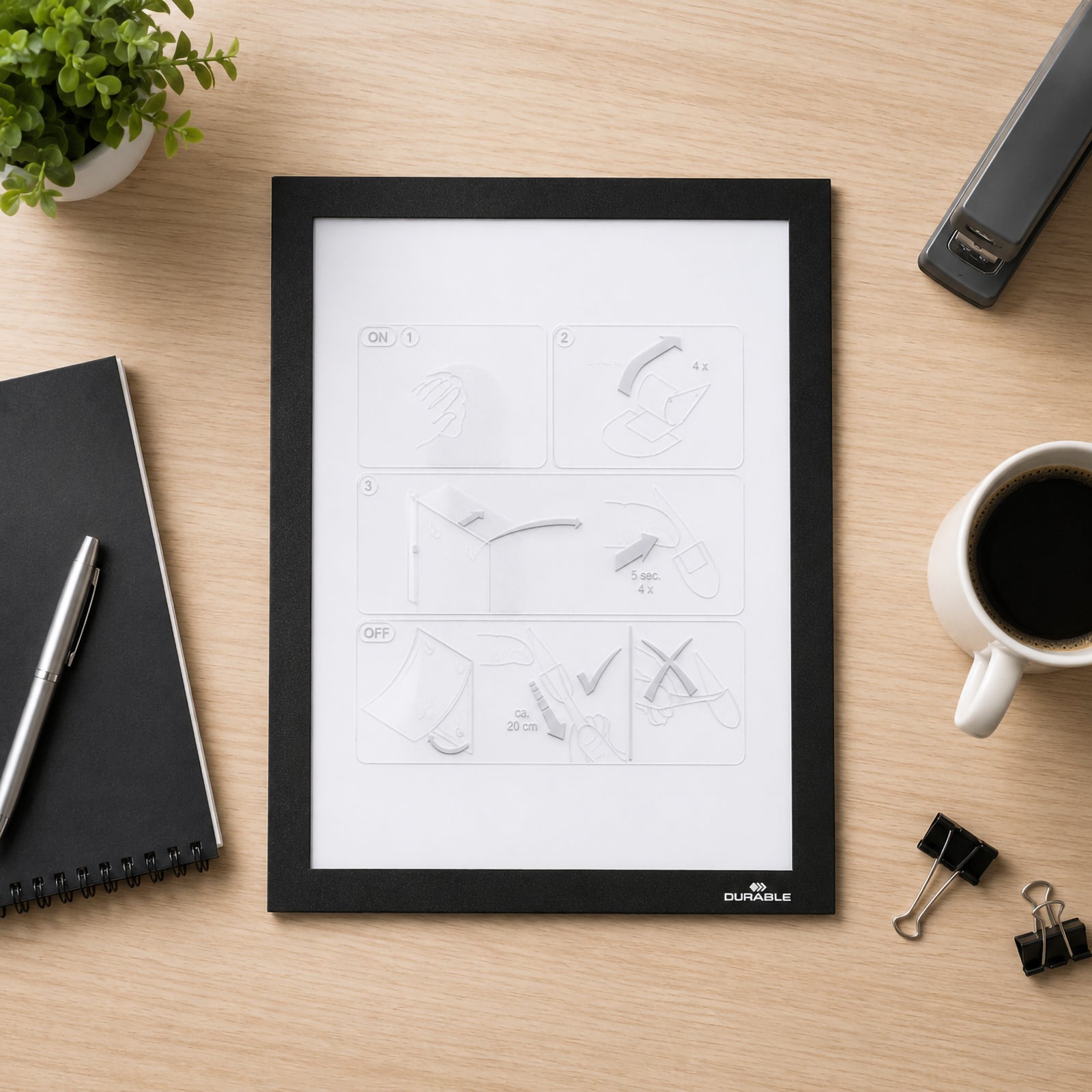 Top view of the Durable DURAFRAME Wallpaper A4 Display Frame placed on a tidy wooden office desk surrounded by a notebook, coffee cup, stapler and stationery. The image highlights the slim black magnetic frame and premium finish before installation.