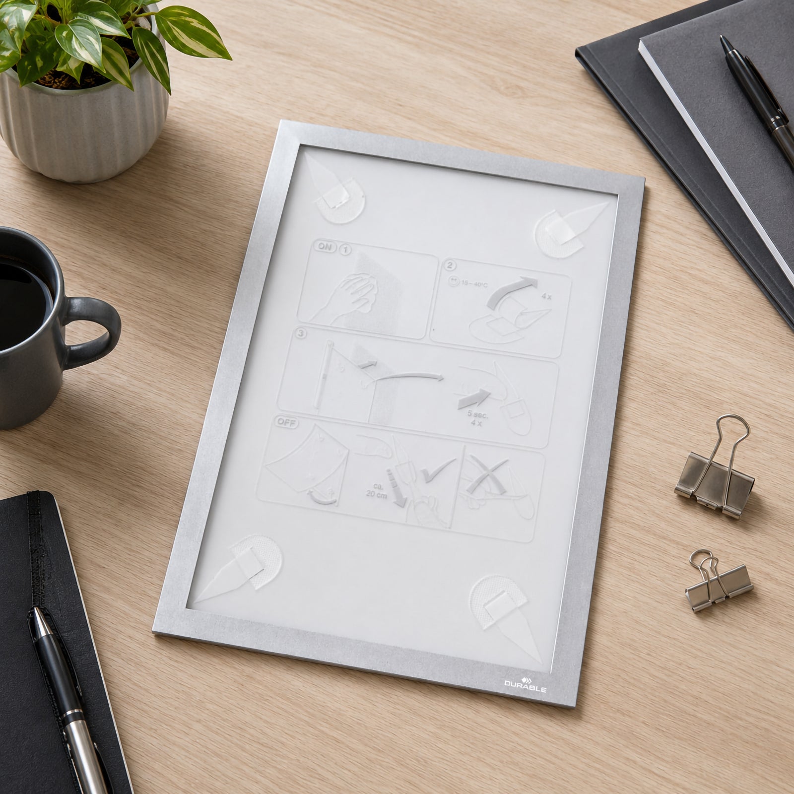 Front view of the Durable Duraframe Wallpaper A4 Silver displayed on a modern wooden desk with adhesive strips visible on the back. A coffee cup, stationery, notebook and binder clips surround the frame, highlighting the easy installation system and reusable adhesive design.