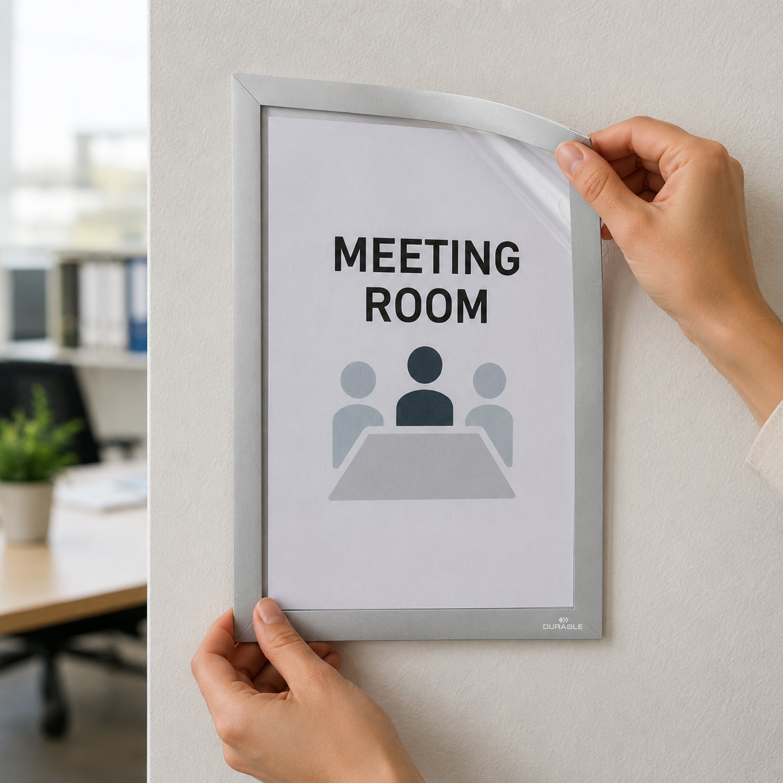 Hands installing the Durable Duraframe Wallpaper A4 Silver onto a painted office wall while lifting the magnetic frame corner to insert a meeting room sign. The image demonstrates quick document replacement and secure mounting on delicate surfaces without damaging wallpaper or painted finishes.