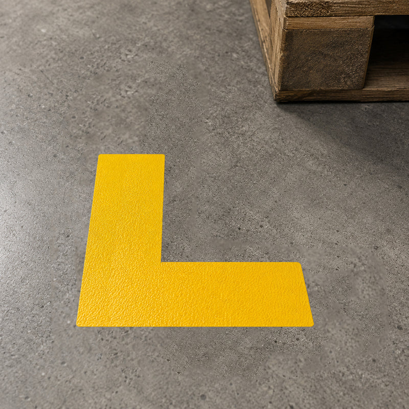 Detailed close up of the Durable Floor Marking Shape L Shape Yellow positioned beside a wooden pallet on an industrial concrete floor. The abrasion resistant floor marker creates a precise ninety degree corner for defining storage locations, walkways or operational zones in warehouses and factories.