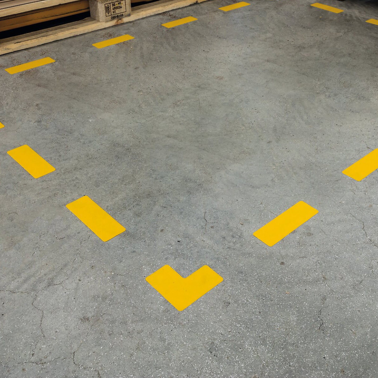 Close up angled view of the Durable Floor Marking Shape L Shape Yellow installed on a concrete warehouse floor. The bright signal yellow L shaped self adhesive floor marker highlights the corner of a designated storage area, demonstrating clear boundary marking for pallets, equipment or work zones.