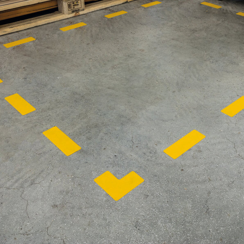 Close up angled view of the Durable Floor Marking Shape L Shape Yellow installed on a concrete warehouse floor. The bright signal yellow L shaped self adhesive floor marker highlights the corner of a designated storage area, demonstrating clear boundary marking for pallets, equipment or work zones.