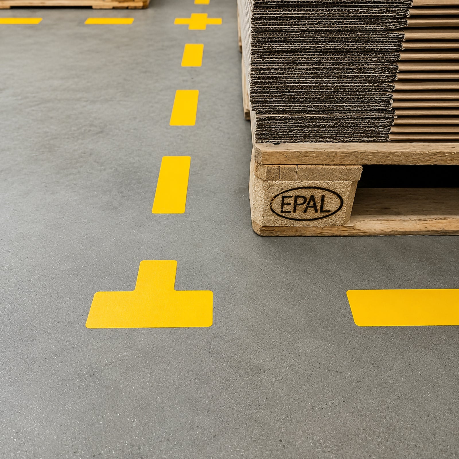 Perspective view of a Durable T Shape Yellow floor marker positioned beside stacked pallets in a warehouse aisle, demonstrating clear boundary marking for organised storage and pedestrian movement.