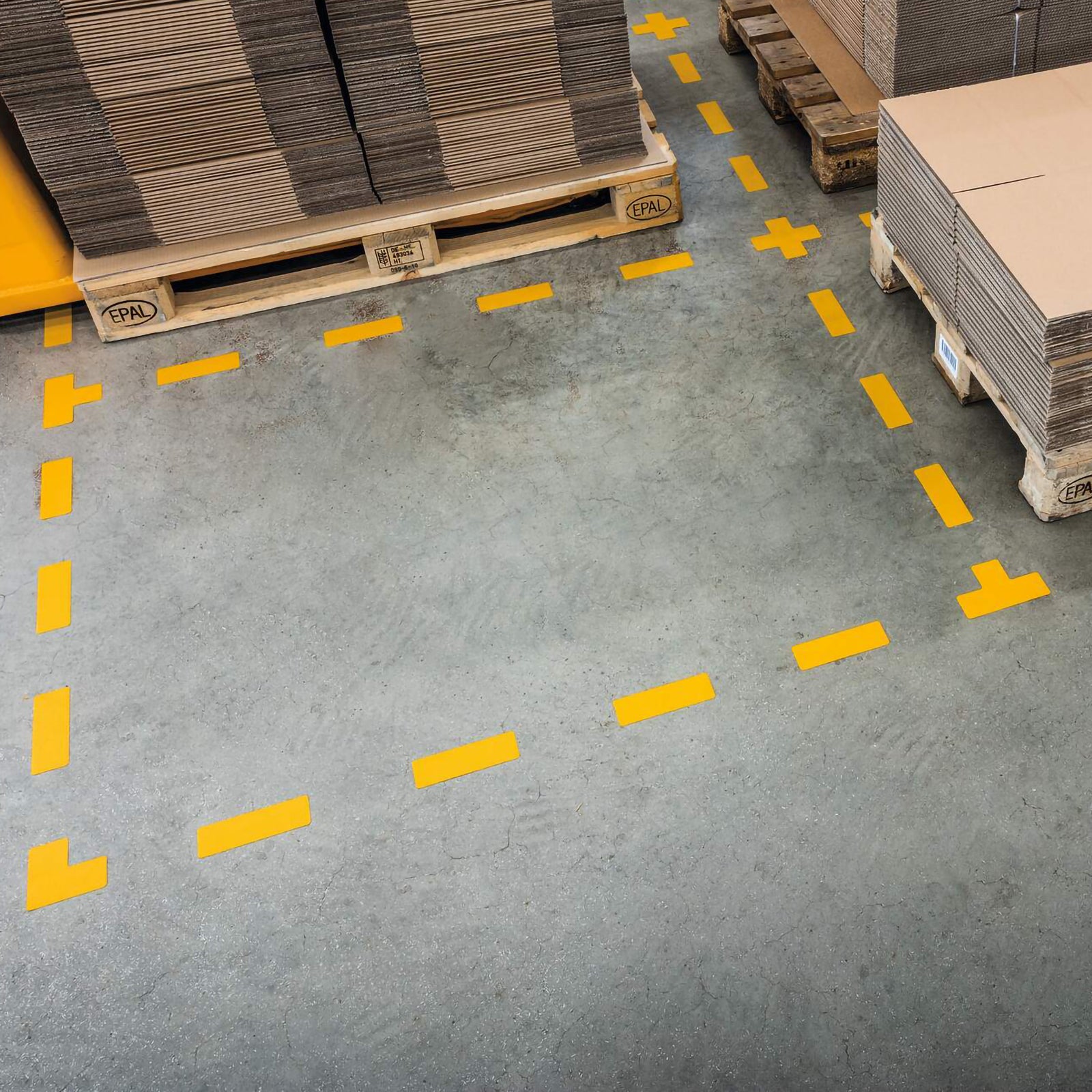 Overhead warehouse scene showing multiple Durable T Shape Yellow floor markers outlining storage bays, pallet locations and traffic routes on a clean industrial concrete floor.