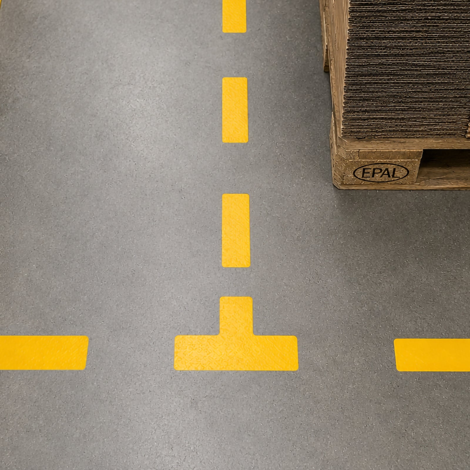 Close up view of a Durable T Shape Yellow floor marker installed at a warehouse aisle intersection beside palletised goods, clearly identifying a traffic junction with bright high visibility floor markings.