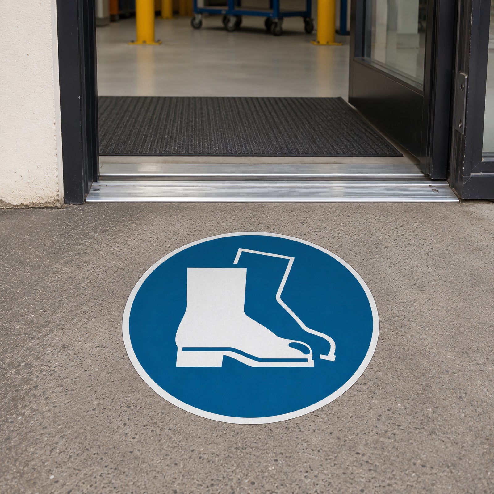 The Durable Floor Marking Sign Use Foot Protection 430mm Blue positioned at the entrance to an industrial facility, clearly identifying a mandatory safety footwear zone before staff enter the workplace.