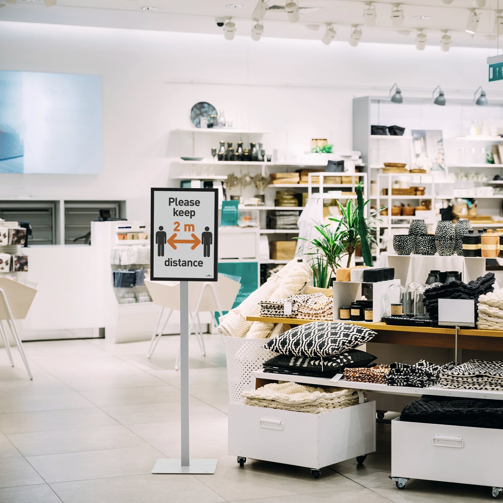 Retail lifestyle scene featuring the Durable Info Stand Floor Basic A3 Silver displaying a social distancing notice within a modern homewares store. The freestanding sign helps communicate customer guidance while blending into contemporary retail interiors.