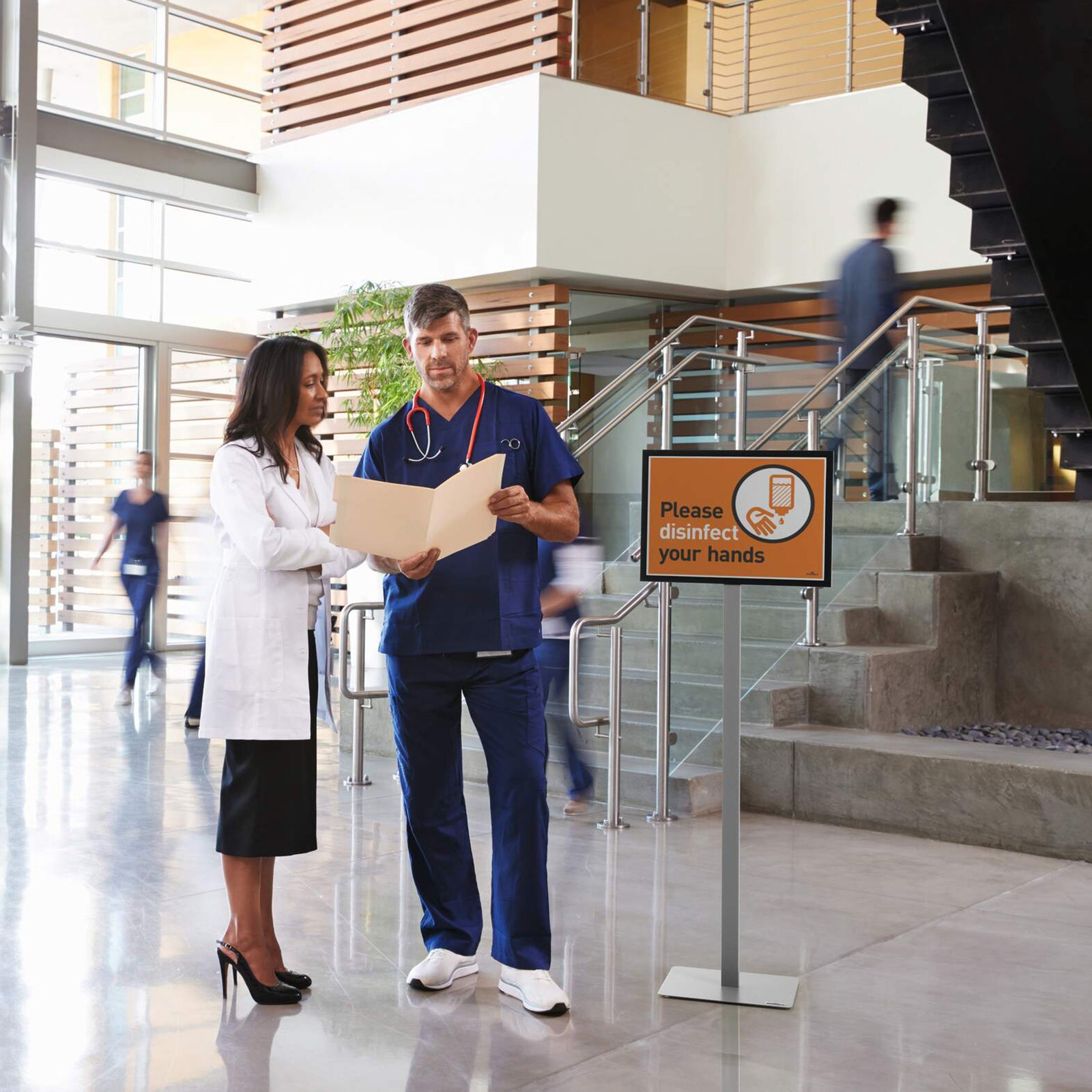 The Durable Info Stand Floor Basic A3 Silver displayed in a hospital lobby presenting a hand hygiene notice while healthcare professionals review patient information nearby. The sturdy floor stand provides clear communication in busy healthcare environments.