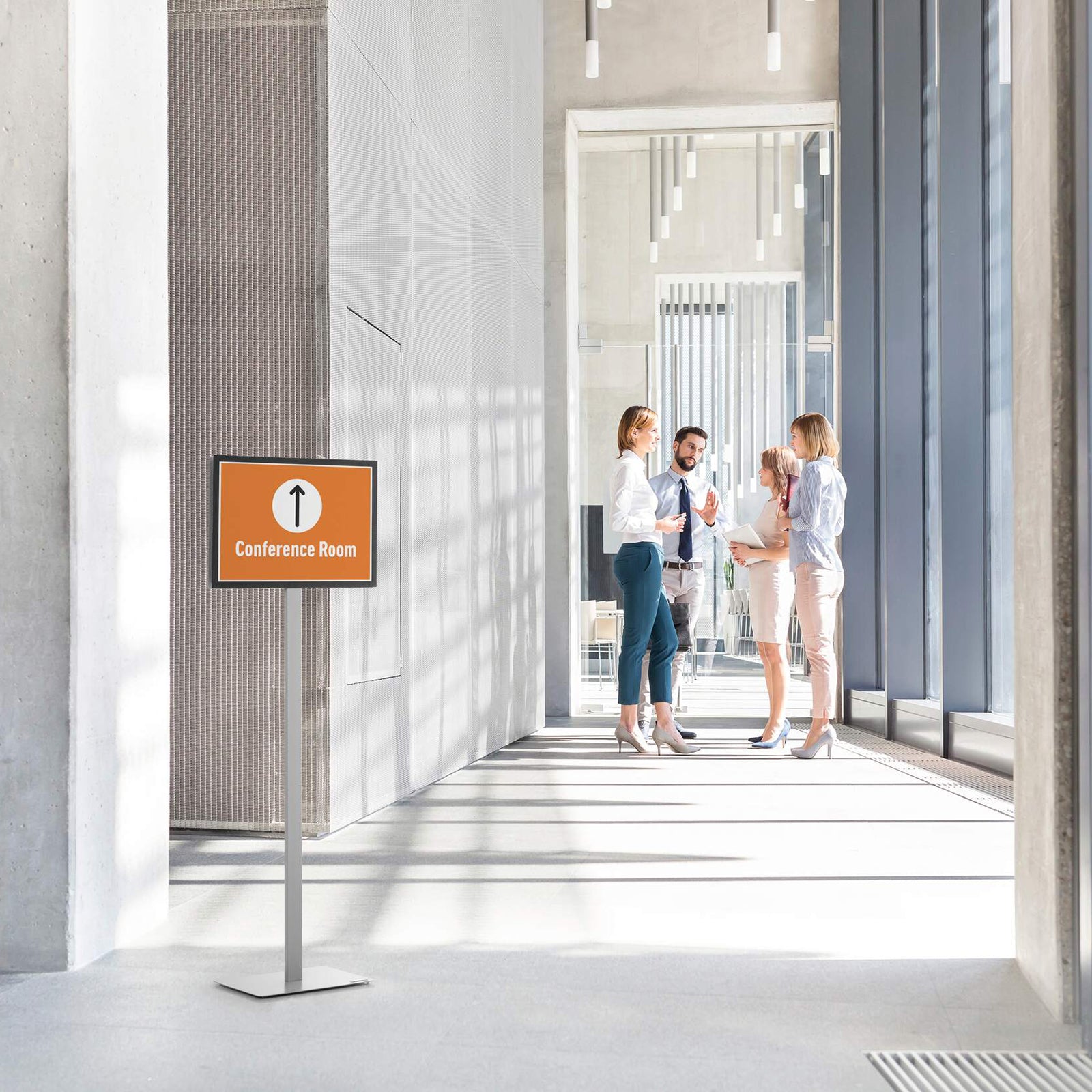 Lifestyle image showing the Durable Info Stand Floor Basic A3 Silver positioned in a bright corporate office corridor displaying a conference room directional sign. The professional double sided information stand helps visitors navigate modern commercial spaces with clear, highly visible signage.