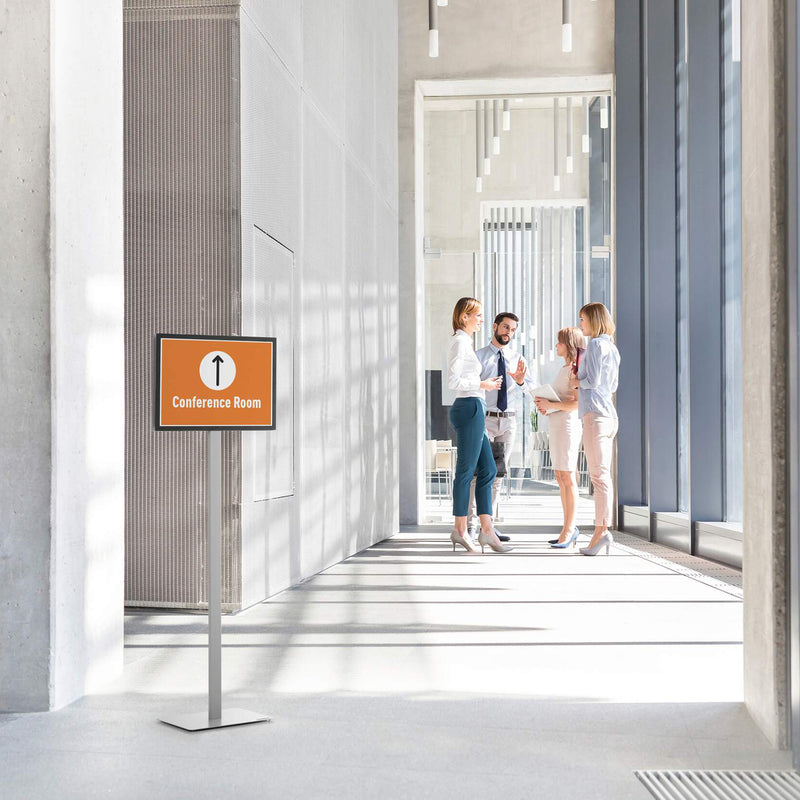 Lifestyle image showing the Durable Info Stand Floor Basic A3 Silver positioned in a bright corporate office corridor displaying a conference room directional sign. The professional double sided information stand helps visitors navigate modern commercial spaces with clear, highly visible signage.