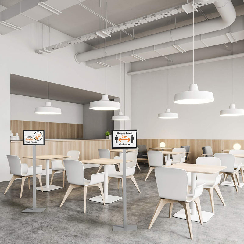 The Durable Info Stand Floor Basic A4 Silver displayed throughout a modern office cafeteria with hygiene and distancing signs positioned between seating areas. The freestanding double sided information displays provide clear visitor guidance while complementing the clean contemporary workspace.