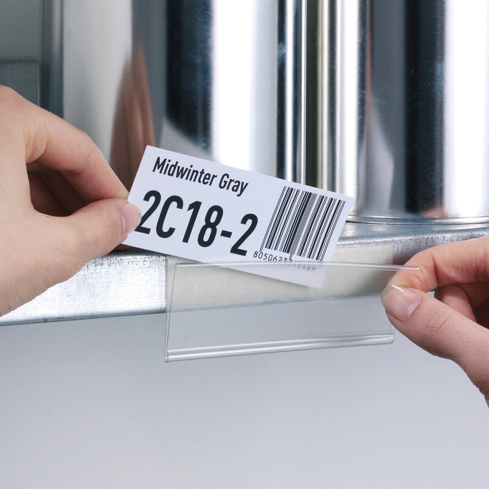 Hands inserting a printed barcode and product identification label into the Durable Scanfix Self Adhesive Label Strip mounted on a metal storage shelf, highlighting the convenient top opening for fast label replacement.