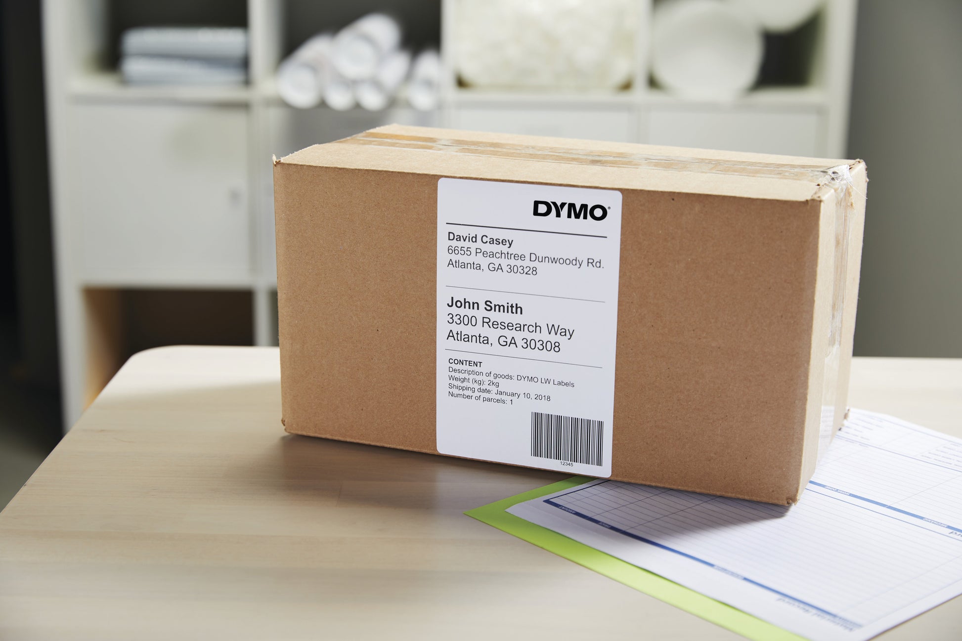 Cardboard carton on a timber desk with a printed Dymo 104 x 159mm shipping label applied, showing sender and recipient addresses and a barcode, shelving in the background.