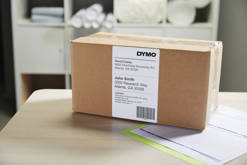 Cardboard carton on a timber desk with a printed Dymo 104 x 159mm shipping label applied, showing sender and recipient addresses and a barcode, shelving in the background.