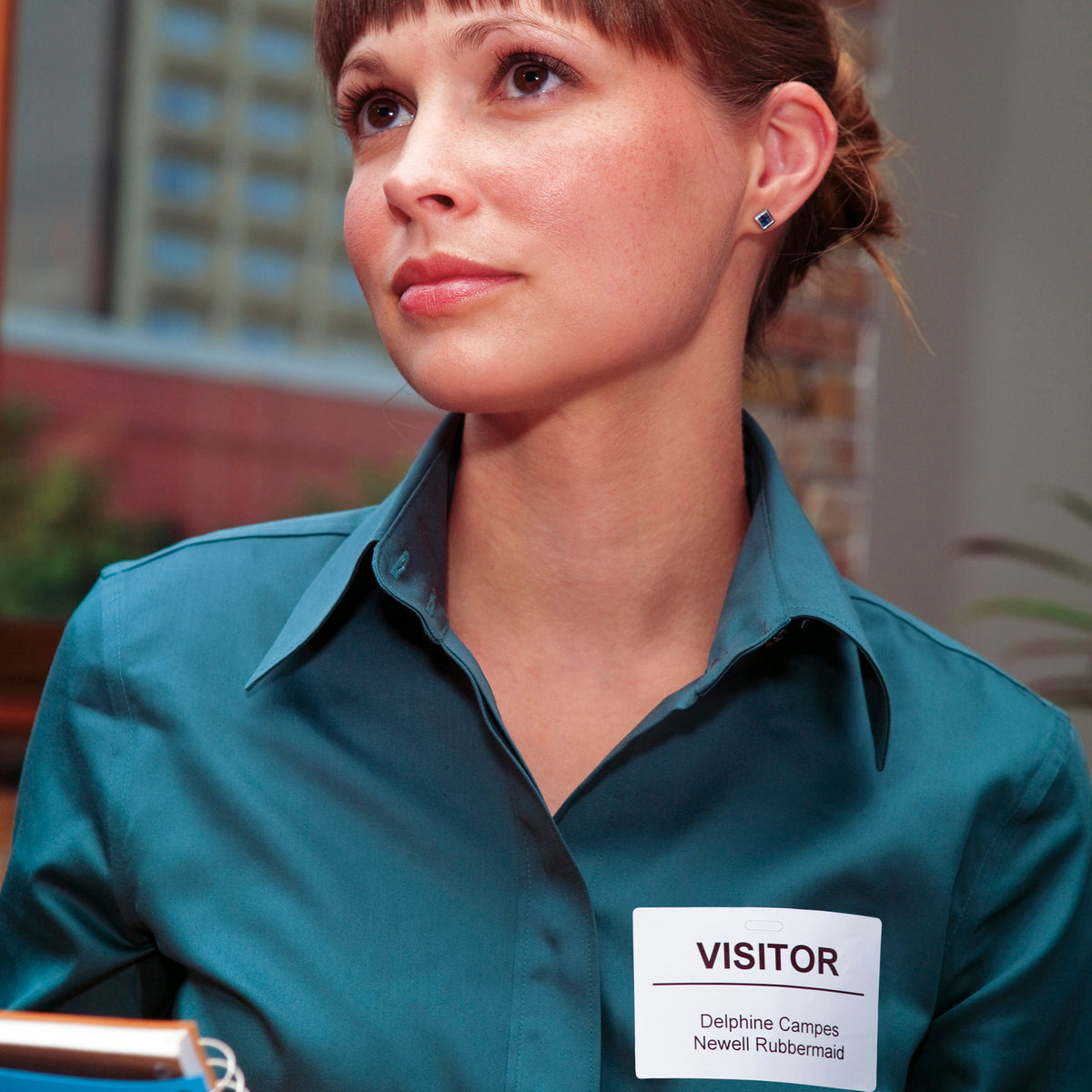 Woman in a teal shirt wearing a white Dymo visitor badge label reading Visitor, Delphine Campos, Newell Rubbermaid.