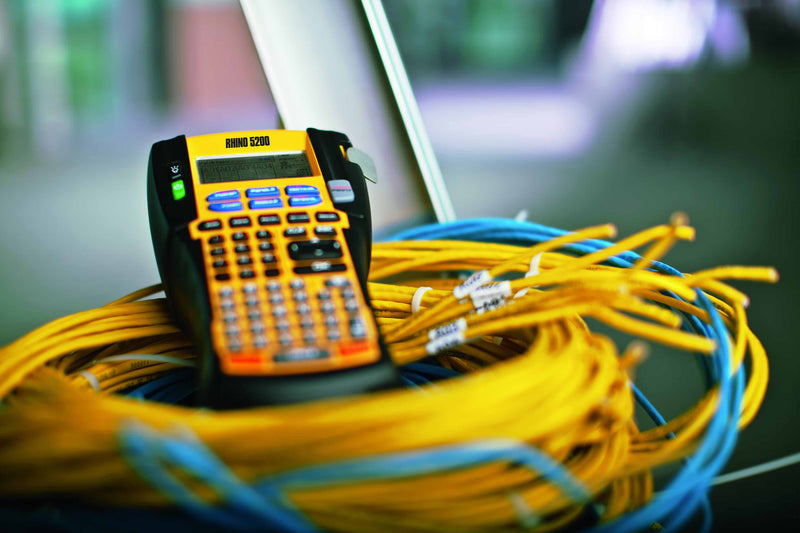 Dymo Rhino label maker with a yellow rubber case sitting on a coil of yellow cable beside blue cables.