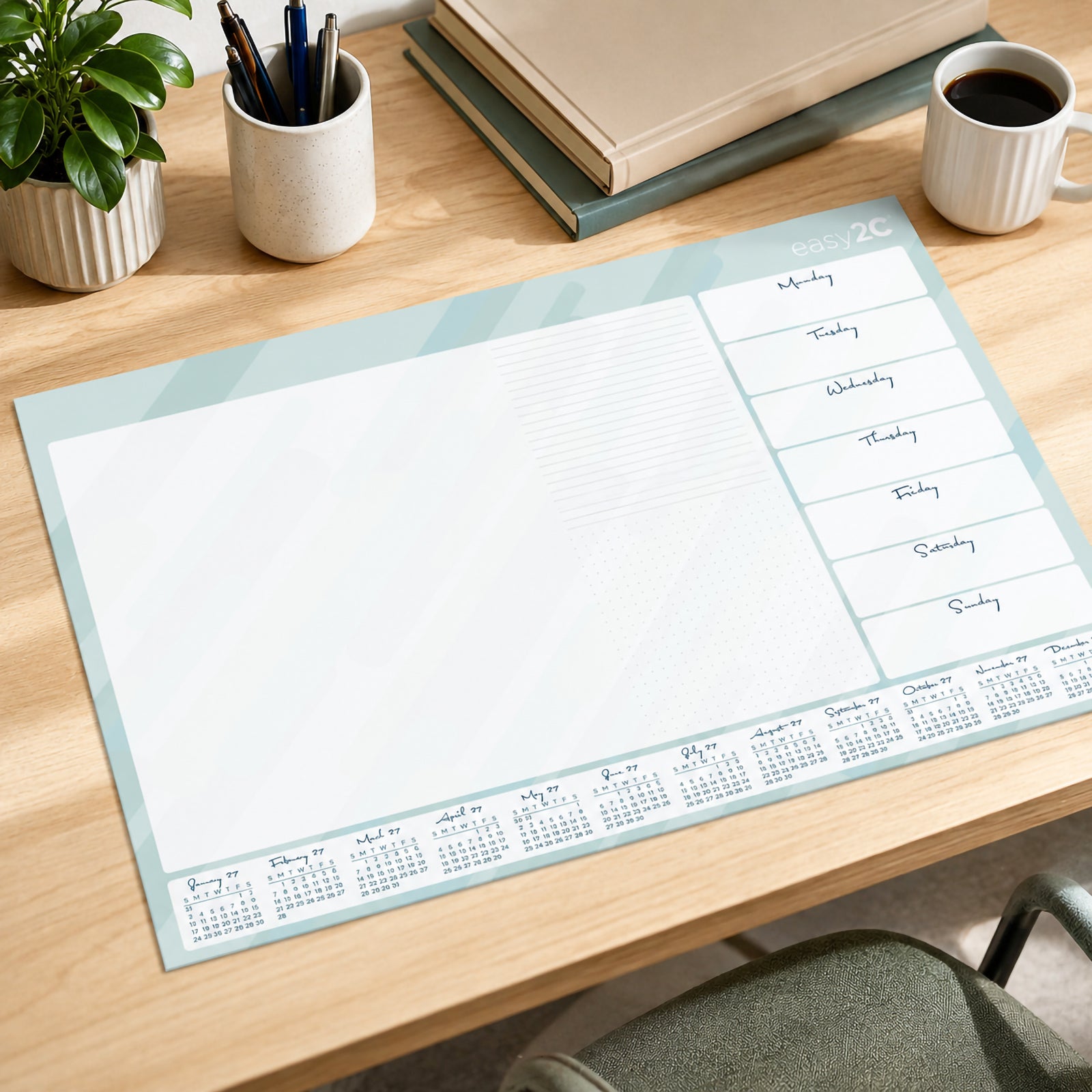 The Easy2C 2027 desk pad displayed on a tidy office desk with coffee, stationery, books and indoor plants. The planner features clearly divided daily sections, note areas and convenient monthly reference calendars along the bottom.