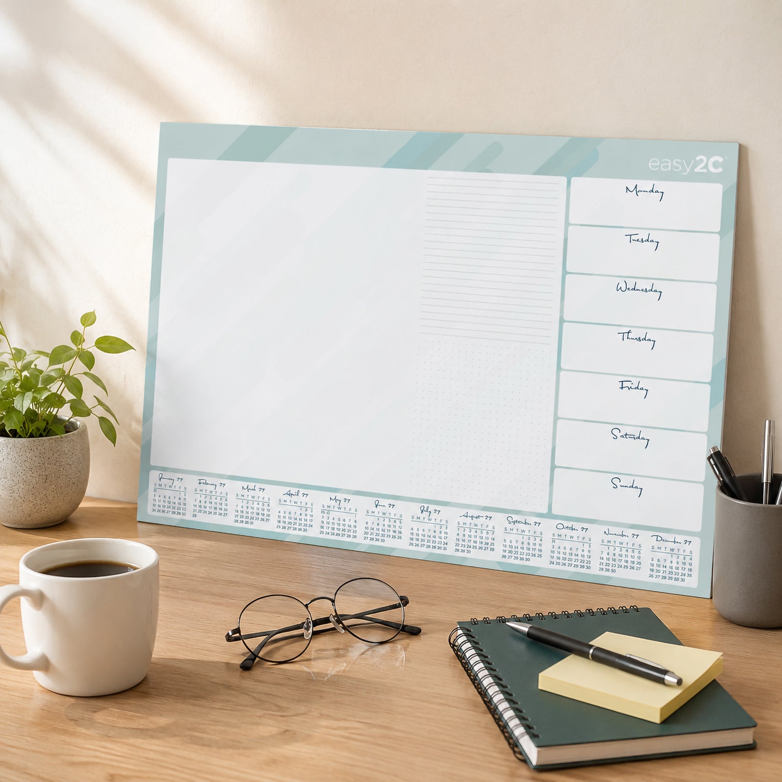 The Easy2C 2027 desk pad leaning against a wall on a modern desk surrounded by notebooks, glasses, pens and coffee. The spacious planning layout demonstrates how the desk pad complements an organised home or office workspace.