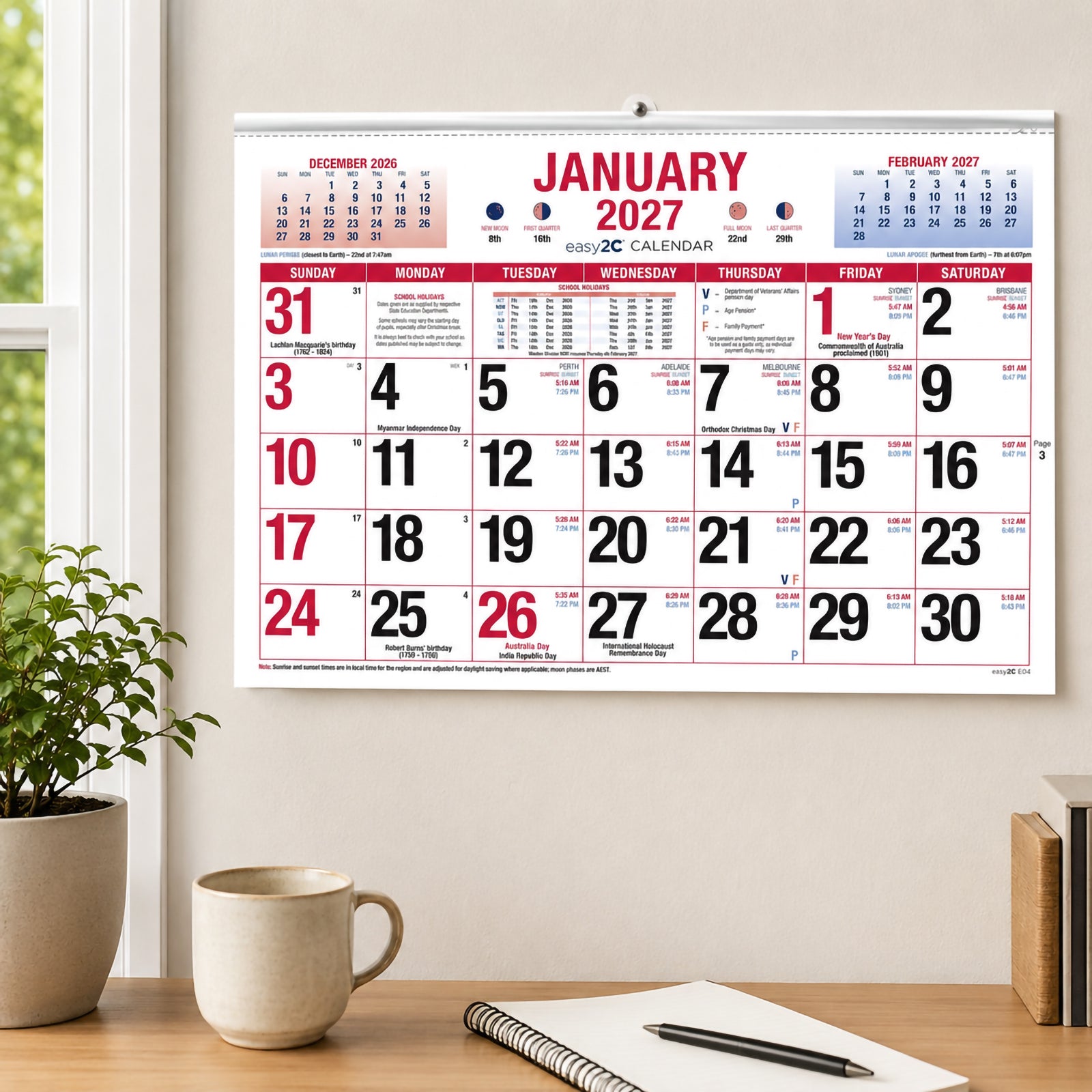 The Easy2C 2027 Australian Date Pad mounted on a clean office wall above a timber desk with a notebook, coffee mug and indoor plant. The large January monthly layout clearly displays spacious date boxes and Australian planning information.