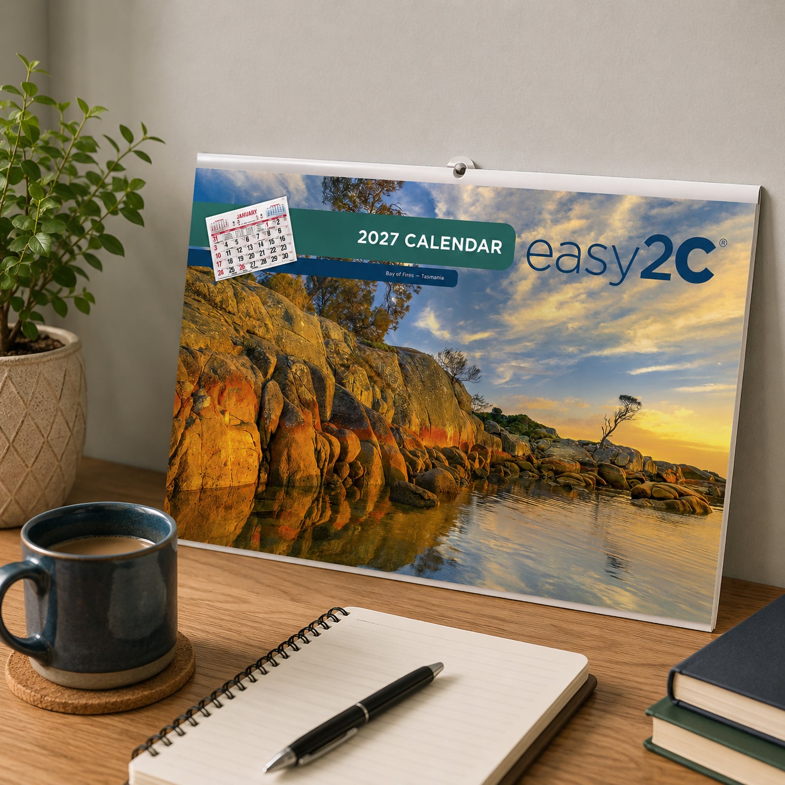 The front cover of the Easy2C 2027 Australian Date Pad displayed on a timber desk in a modern workspace. The cover showcases the scenic Bay of Fires in Tasmania alongside Easy2C branding and the 2027 calendar title.