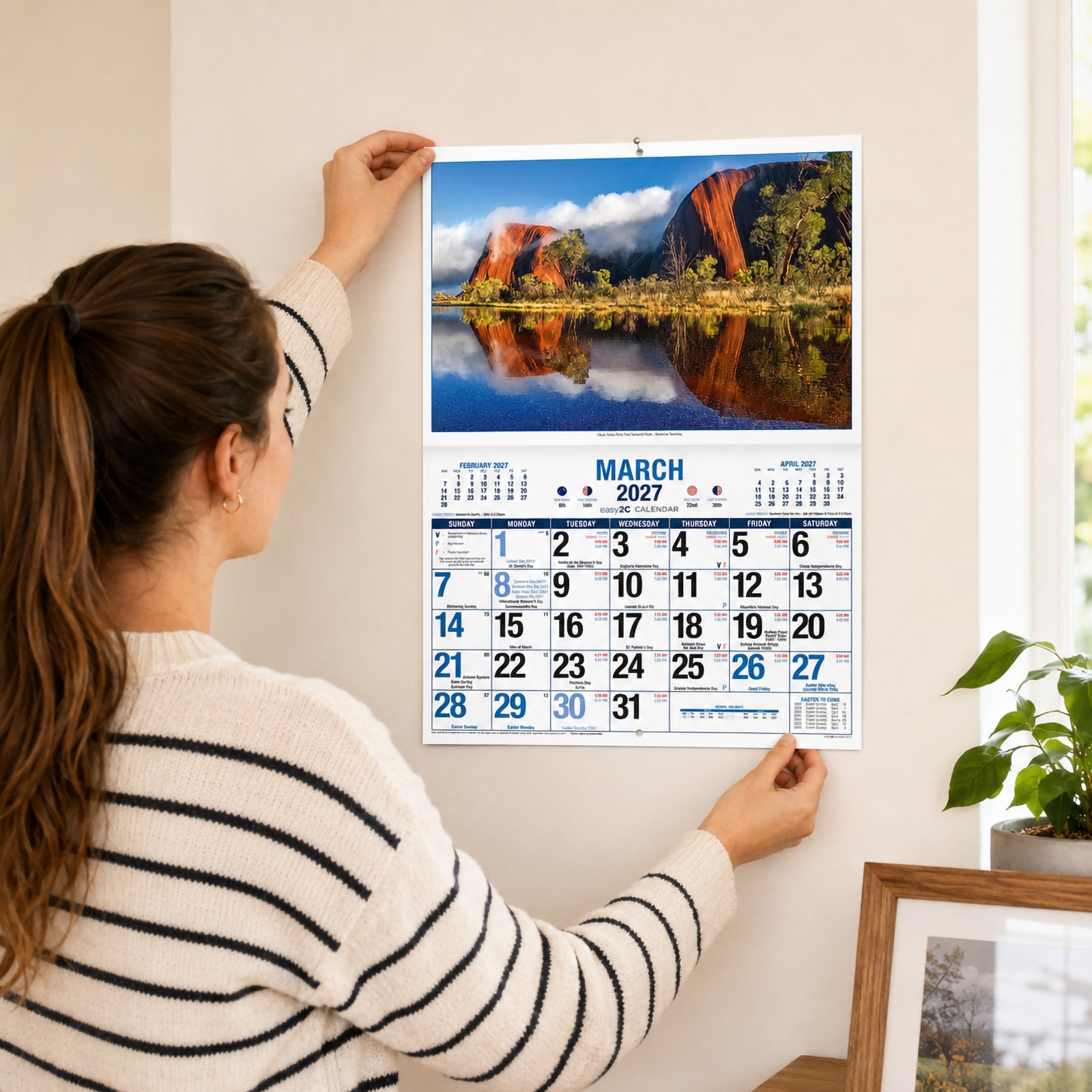 A person hangs the Easy2C Calendar 2027 Stitched A4 on a light coloured wall in a bright modern home. The March page features Uluru reflected in calm water with a spacious monthly planner beneath, demonstrating the calendar in everyday use.