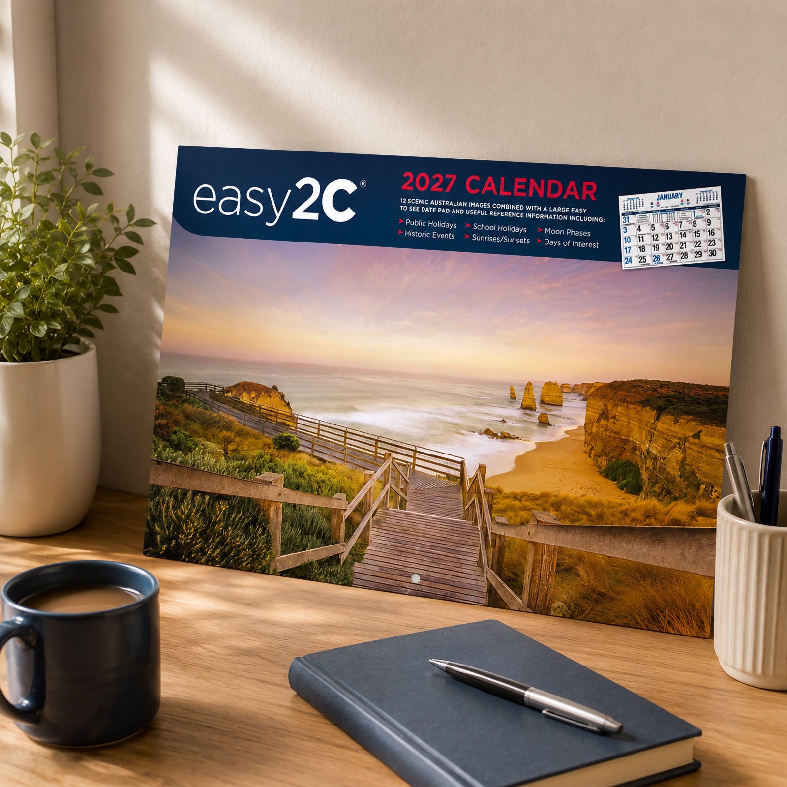 The Easy2C Calendar 2027 Stitched A4 cover leaning against a wall on a timber desk beside a notebook, coffee mug and stationery. The cover features the iconic Twelve Apostles coastline at sunset, creating an attractive office display.