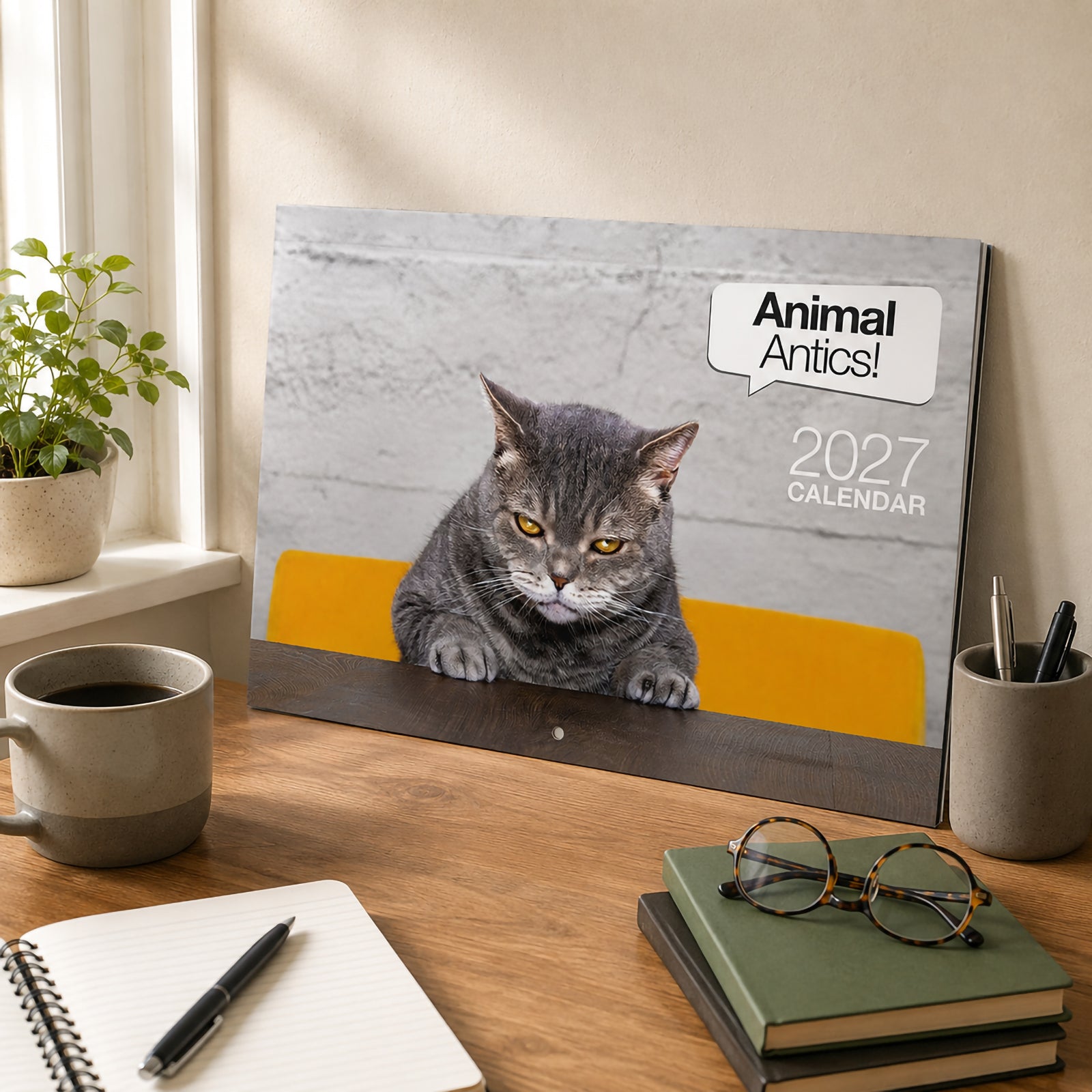 The front cover of the Easy2C Animal Antics 2027 calendar leaning against a wall on a wooden desk. Office accessories including a notebook, glasses, coffee cup and pen create a welcoming workspace while highlighting the amusing cat themed cover design.