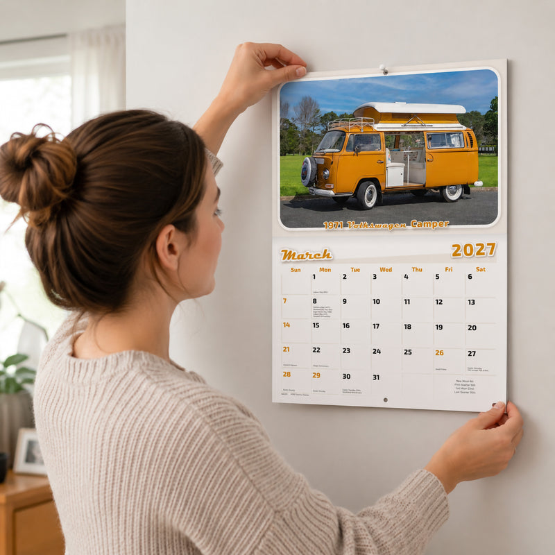 Easy2C Calendar 2027 Stitched Custom Classics A4 mounted on a wall as a person hangs it in a bright modern home. The March page displays a vibrant yellow 1971 Volkswagen Camper with a spacious monthly planner beneath, demonstrating the calendar in everyday use.