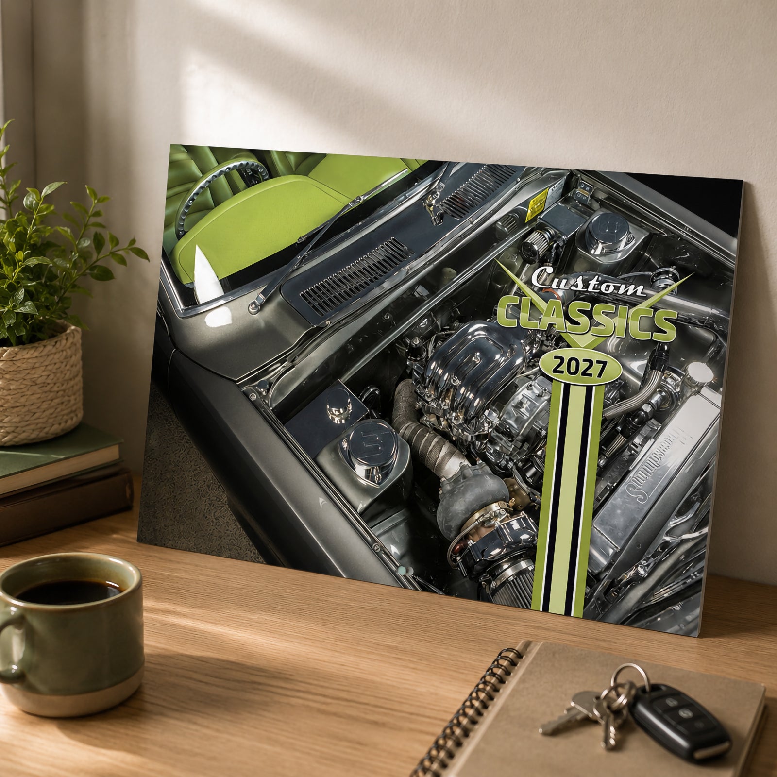 Easy2C Calendar 2027 Stitched Custom Classics A4 front cover standing on a wooden desk beside a coffee cup, notebook and keys. The cover artwork showcases a detailed polished classic car engine with bold retro Custom Classics 2027 branding.