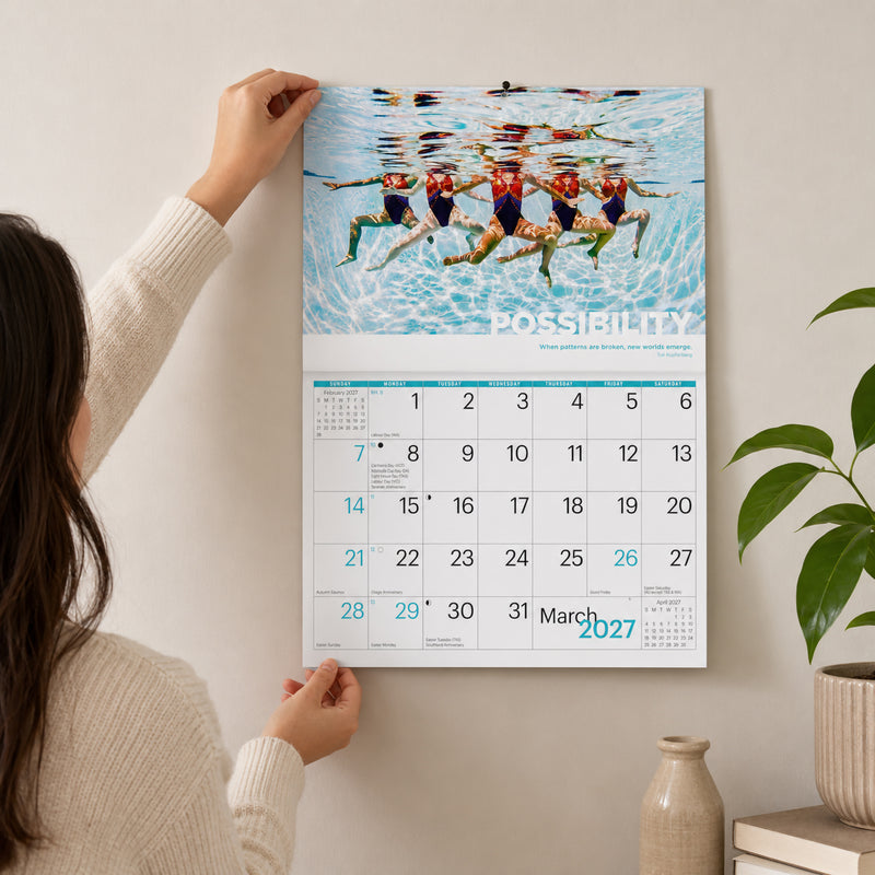 Easy2C Inspirations 2027 stitched A4 wall calendar being mounted on a light coloured wall by a user. The March monthly page displays an underwater synchronised swimming photograph with a motivational quote, demonstrating the practical calendar layout and compact wall mounted design.