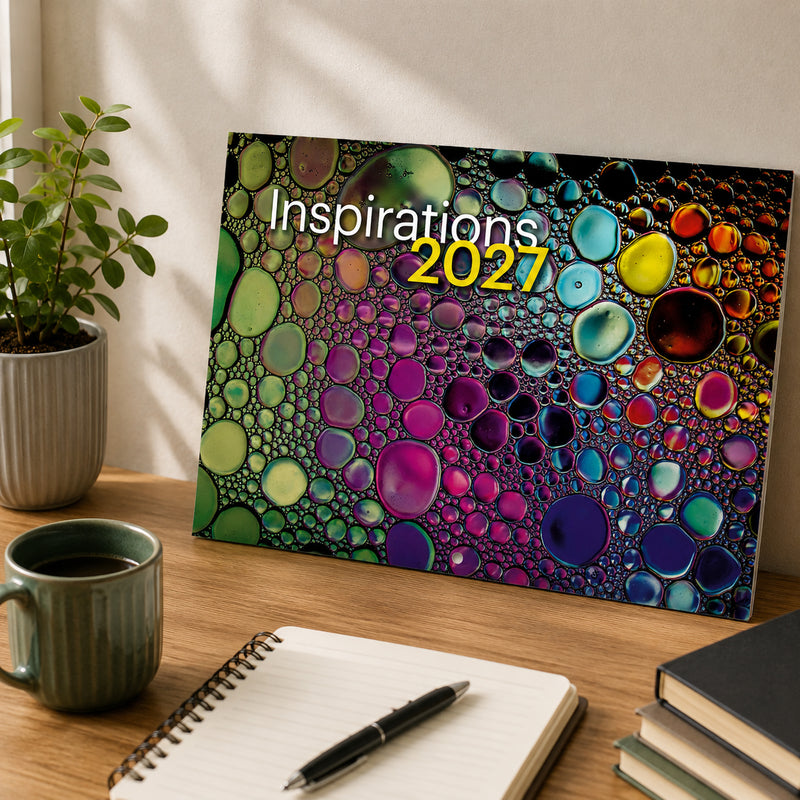 Front cover of the Easy2C Inspirations 2027 stitched A4 calendar resting on a timber desk in a contemporary office. The cover features vibrant abstract bubble artwork beside a notebook, coffee mug and indoor plant, highlighting the colourful design.