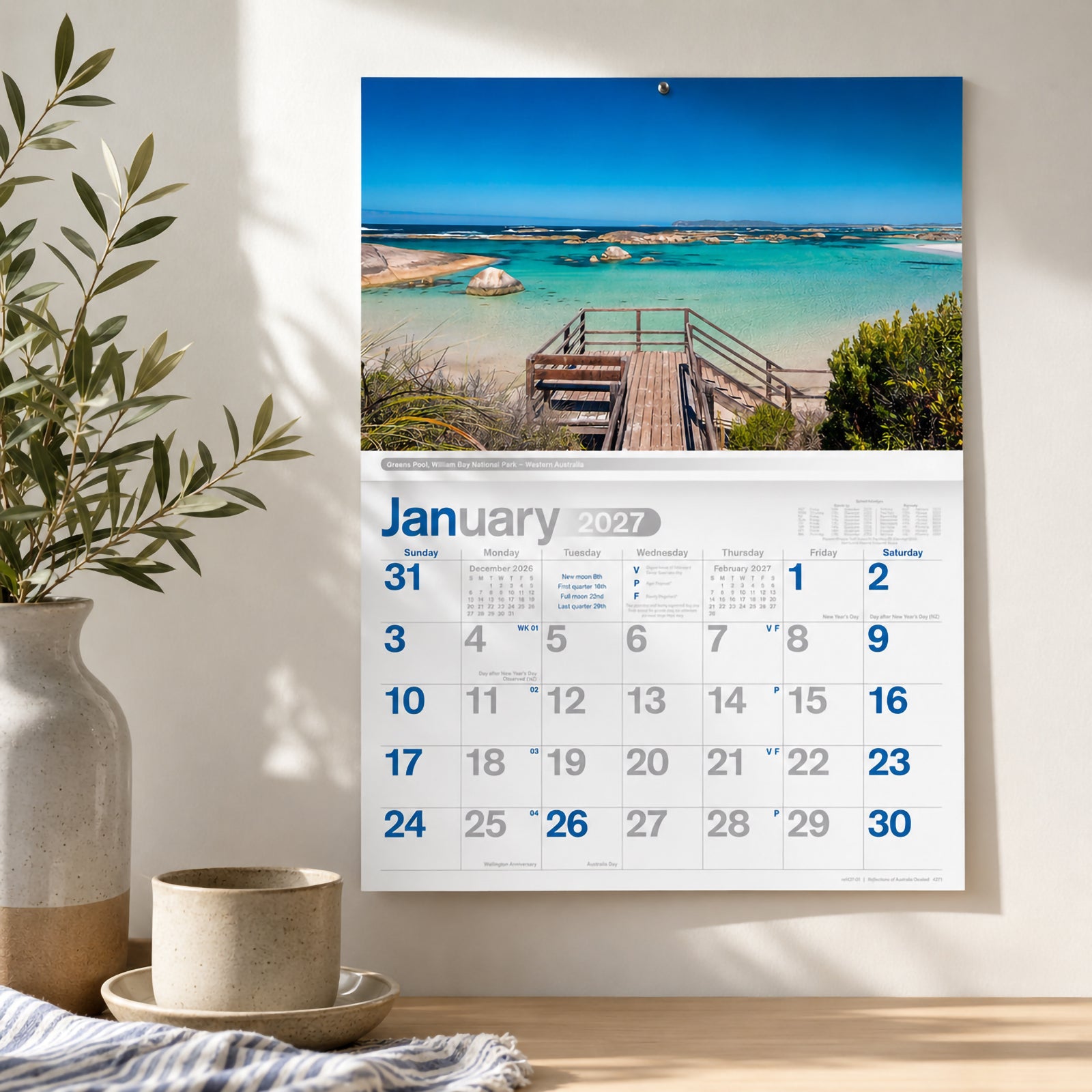The Easy2C Reflections of Australia 2027 A4 stitched wall calendar displayed on a bright interior wall featuring the January page. The scenic Greens Pool photograph overlooks crystal clear turquoise waters and a timber viewing platform, highlighting the calendar's vivid photography and clean monthly layout.