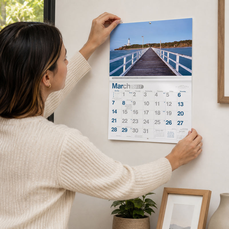 A lifestyle scene showing the Easy2C 2027 Reflections of Australia stitched A4 wall calendar being hung on a modern wall by a person. The March page displays the Point Lonsdale Pier and Lighthouse in Victoria, demonstrating the calendar's practical size and elegant presentation within a contemporary home environment.