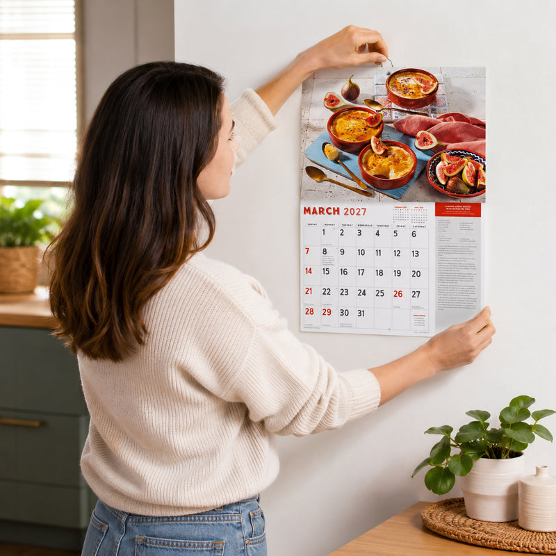 Lifestyle image showing a person hanging the Easy2C 2027 Sugar and Spice stitched A4 calendar on a kitchen wall. The March recipe page displays almond creme brulee with marinated figs, demonstrating the practical wall hanging format in a bright modern home.