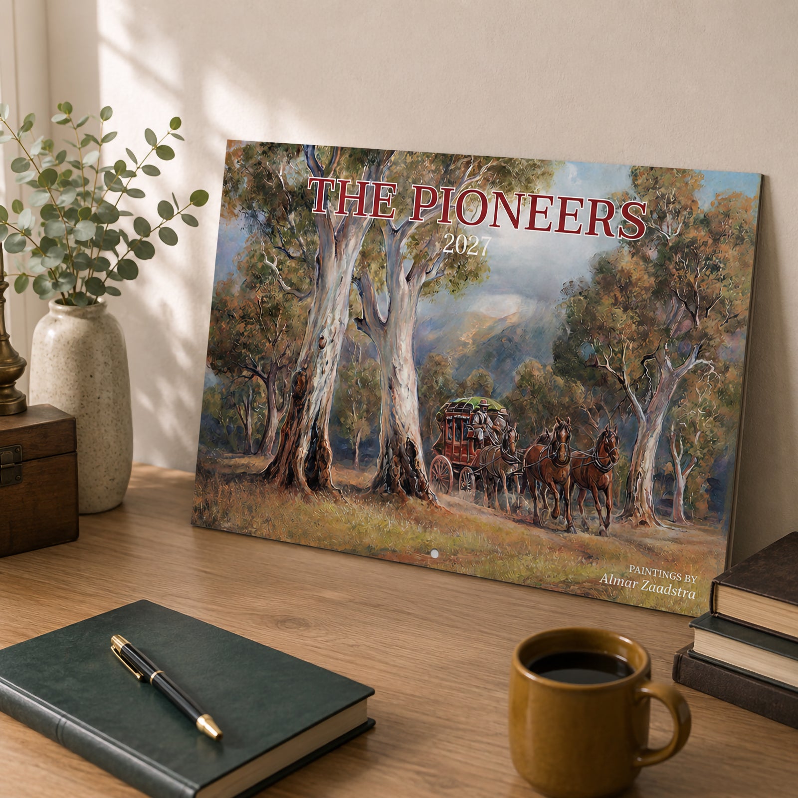 Front cover of the Easy2C 2027 The Pioneers stitched calendar leaning on a timber desk with books, plant and coffee cup, showcasing the beautiful historical landscape artwork by Almar Zaadstra.