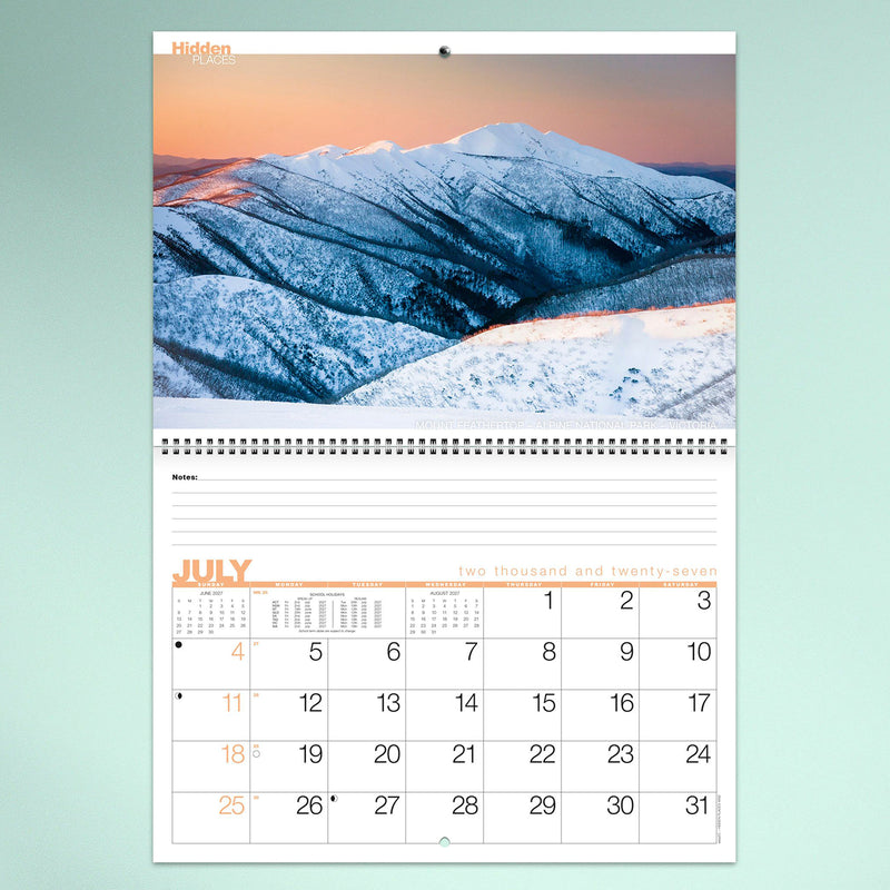 The July calendar page featuring snow covered alpine mountains in Victoria. Crisp winter scenery contrasts beautifully with the practical calendar grid and lined notes area.