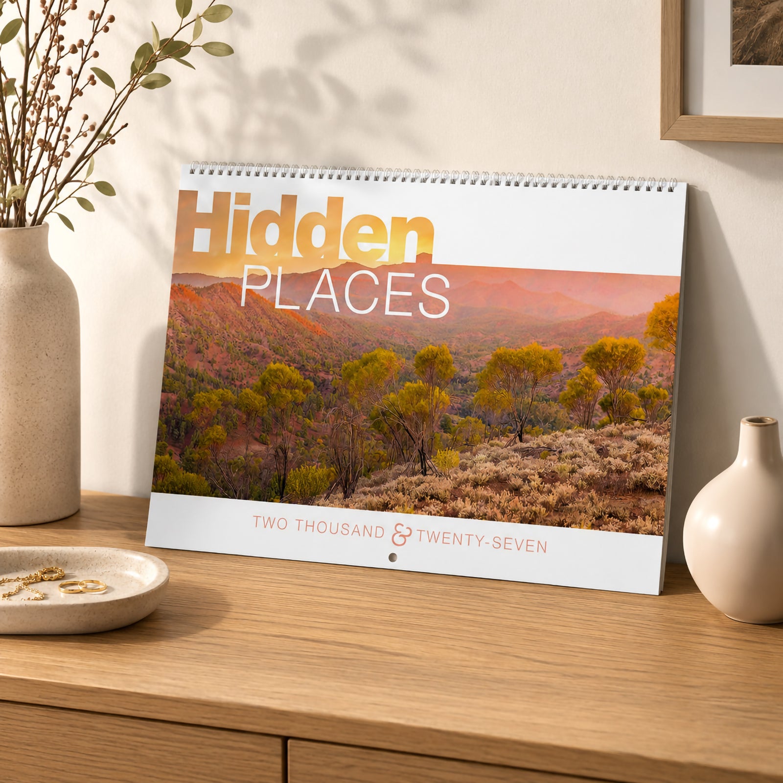 The Hidden Places 2027 calendar cover resting on a timber cabinet in a contemporary interior. The cover showcases warm Australian outback scenery with premium wiro binding and elegant typography, creating a stylish display before opening.