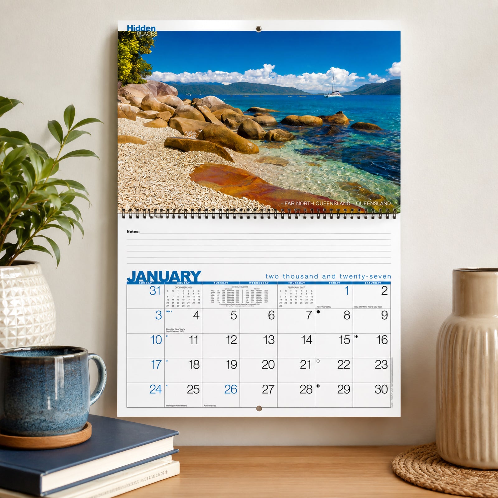 The Easy2C Hidden Places 2027 wall calendar displayed on a wall above a timber desk with indoor plants, books and decorative accessories. The January page features a vibrant Australian coastal scene with generous monthly planning space below.