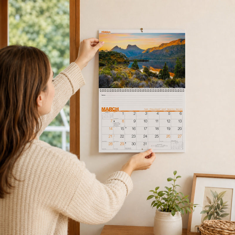 A lifestyle image showing a person hanging the Easy2C Hidden Places 2027 wall calendar on a light coloured wall inside a modern home. The calendar displays a scenic mountain landscape with a spacious monthly planner beneath, highlighting its practical size and attractive presentation.