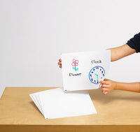 Child holding up a plain white Educational Colours dry erase board with a drawn flower and clock, with more blank boards stacked on a wooden table