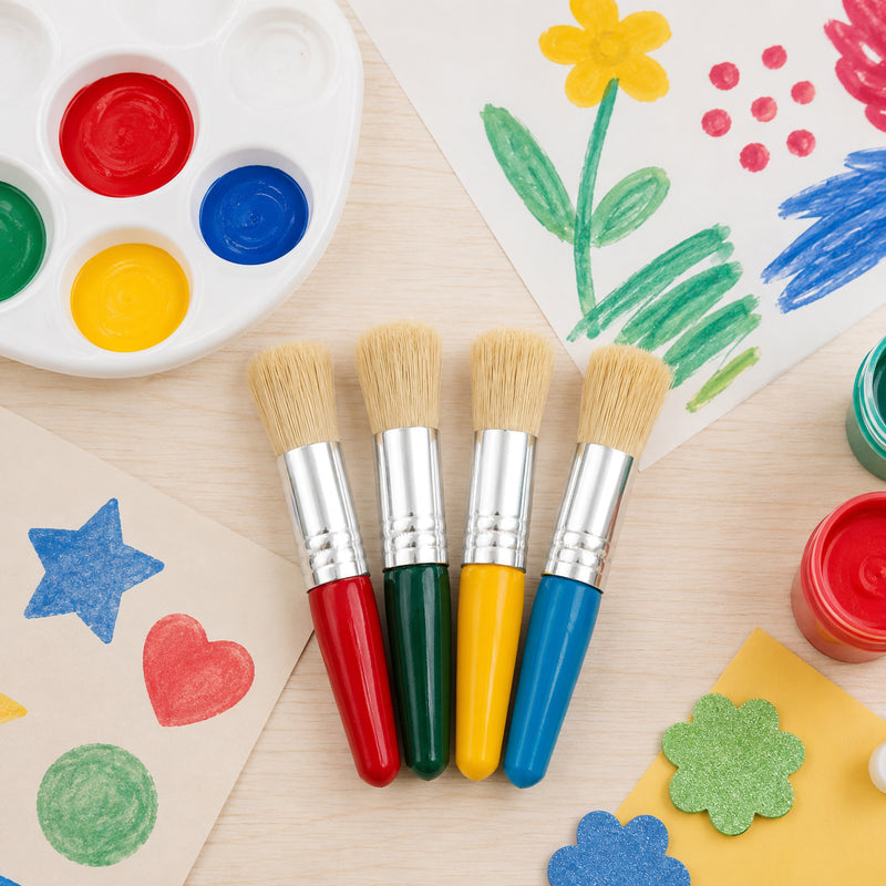 Top view of four Educational Colours Baby Stubby Brushes with assorted red, green, yellow and blue handles arranged between colourful children's artwork, a paint palette and craft shapes. The creative classroom setting highlights the short 100mm brush format and broad bristles suited to young children's painting projects.
