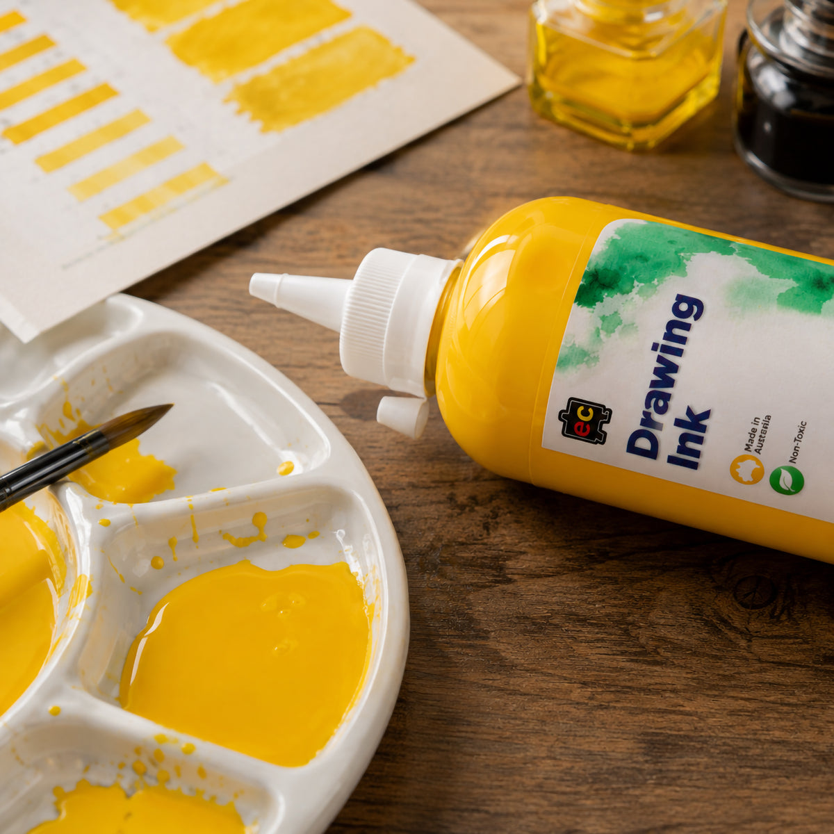 Close up view of Educational Colours Drawing Ink 500ml Brilliant Yellow positioned beside a paint palette filled with vivid yellow ink. Brushes, yellow colour swatches and small ink containers complete the studio setting, highlighting the dispensing bottle and rich colour for painting, illustration and creative experimentation.