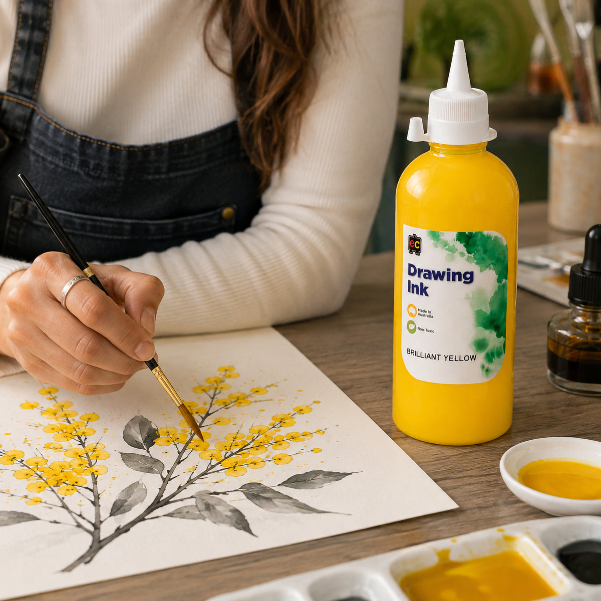 Artist applying Educational Colours Brilliant Yellow Drawing Ink with a fine brush to a detailed botanical artwork featuring yellow blossoms and dark leaves. The 500ml bottle stands prominently on the timber worktable beside palettes and art materials, demonstrating the vibrant ink in use for controlled painting and illustrative details.