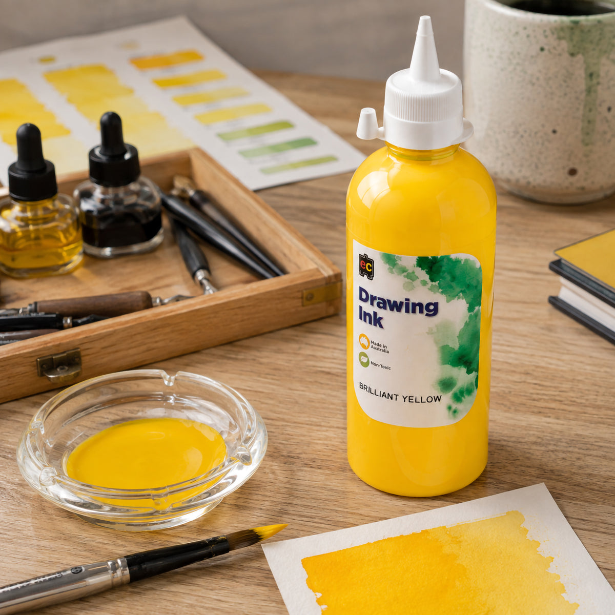 Educational Colours Brilliant Yellow Drawing Ink 500ml displayed upright on an organised artist workspace. A dish of yellow ink, loaded paintbrush, colour swatches and drawing tools surround the bottle, showcasing the bright yellow tone and its suitability for expressive washes, detailed brushwork and mixed media art.
