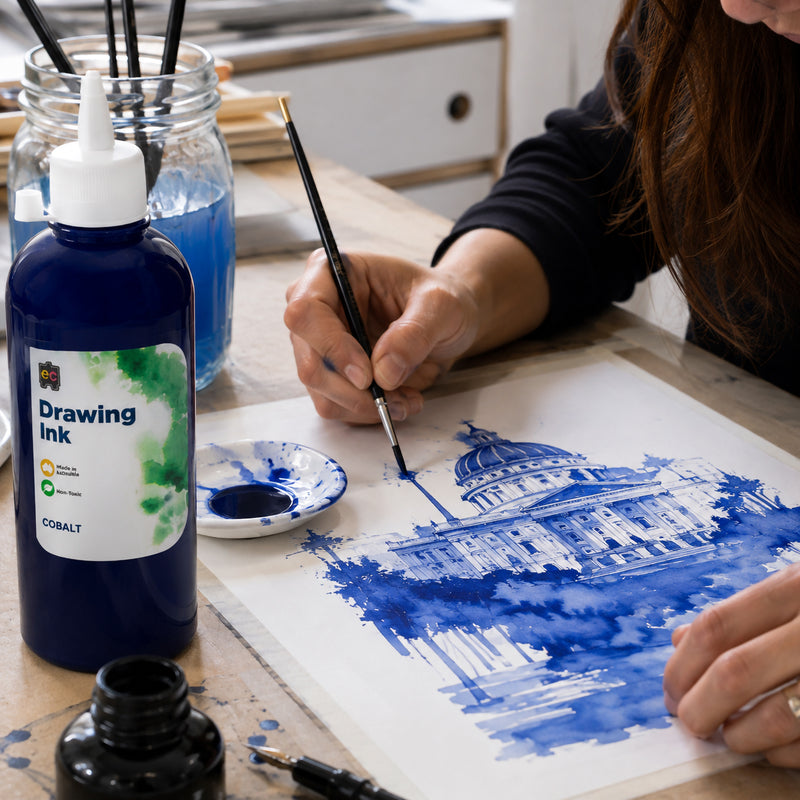 Artist using a fine brush with Educational Colours Drawing Ink 500ml Cobalt to create an intricate blue architectural illustration of a domed building. The Cobalt bottle, ink dish and painting tools are arranged around the artwork, demonstrating detailed brush control and expressive blue washes in a working studio environment.