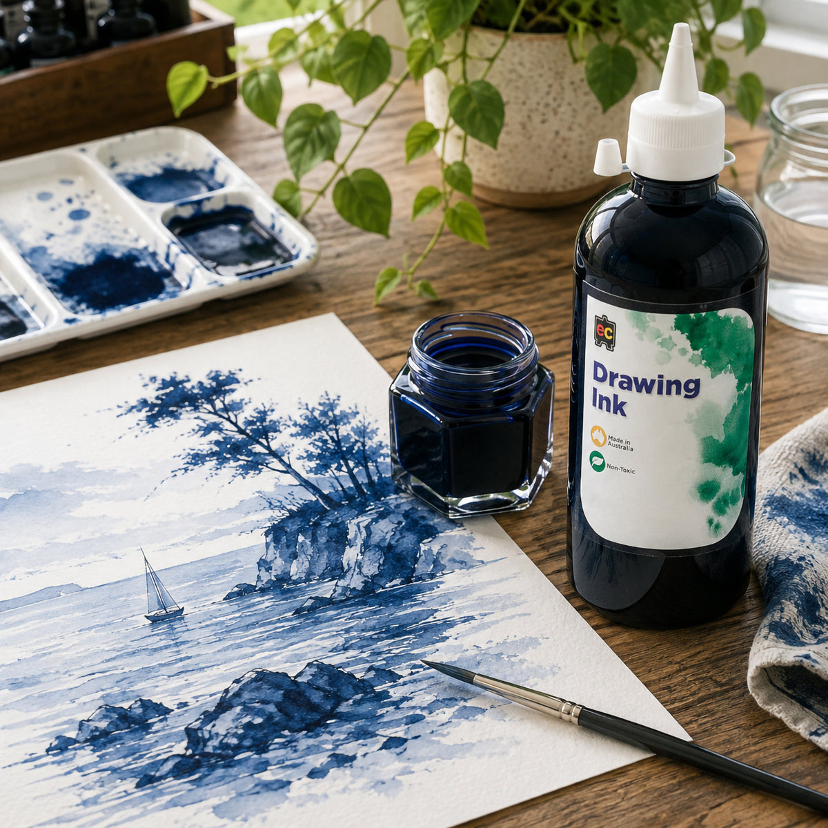 Educational Colours Drawing Ink 500ml Prussian Blue displayed upright beside a completed coastal artwork featuring rocky formations, trees, water and a small sailing boat. The studio arrangement includes a fine brush, palette and ink jar, showcasing deep blue details alongside softer diluted washes.