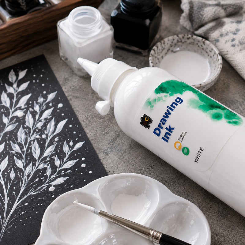 Close up creative workspace featuring Educational Colours Drawing Ink 500ml White beside a black card decorated with detailed white botanical branches. A paint palette, fine brush and white ink dishes surround the bottle, highlighting the strong visual contrast achievable when using white drawing ink on dark surfaces.