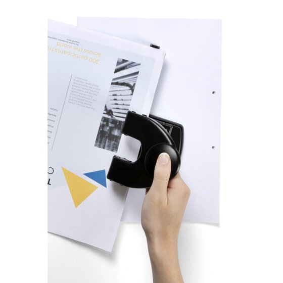 Hand pressing the round button of a black Esselte D25 punch as paper is fed in, with a punched sheet behind